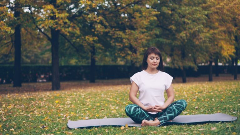 Pretty young woman is relaxing sitting in lotus pose on yoga mat on grass in park and breathing resting after individual practice. Meditation and nature concept.